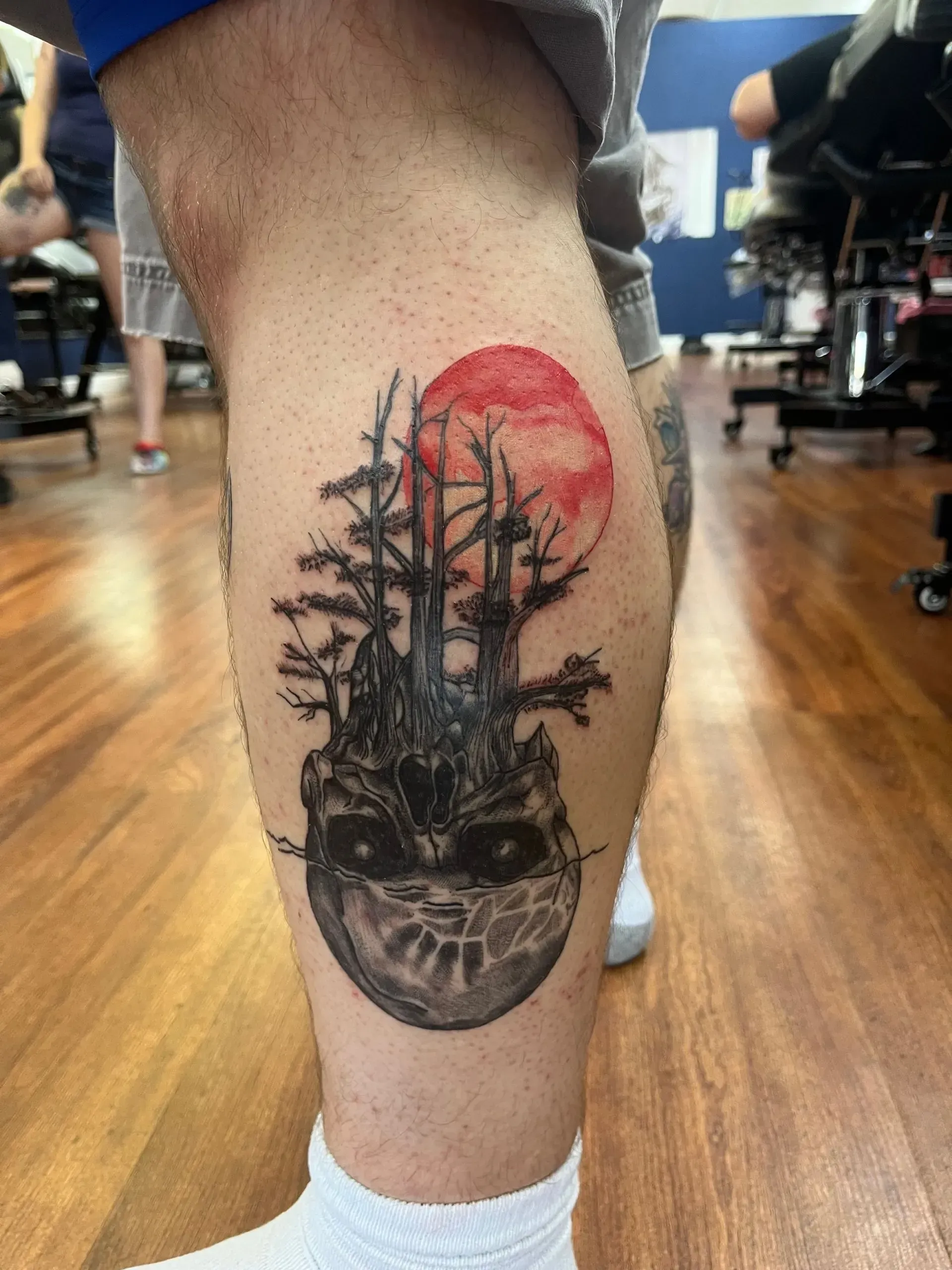 Calf tattoo of a skull with pine trees and a red sun, on a leg in a gym setting
