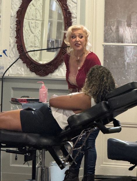 Woman in red top speaking beside a person reclining in a salon chair with a mirror behind them