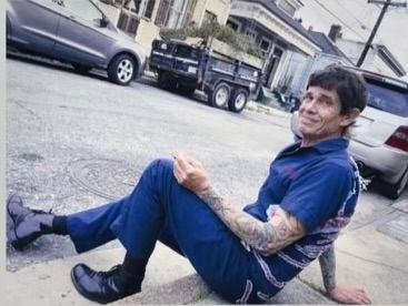 Person sitting on a street curb beside parked cars, wearing blue clothes and smiling at the camera.