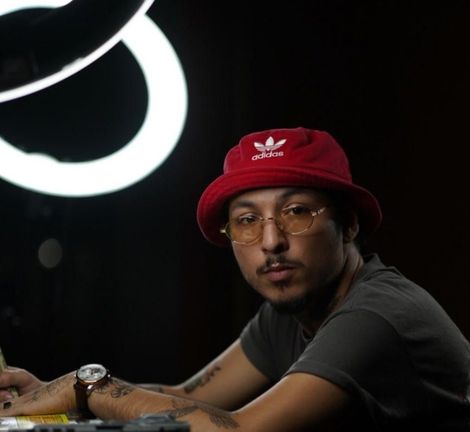 Man in a red bucket hat and glasses sits at a desk against a dark background with a bright ring light.