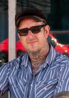 Man in sunglasses and a cap, wearing a blue striped shirt, standing outdoors.