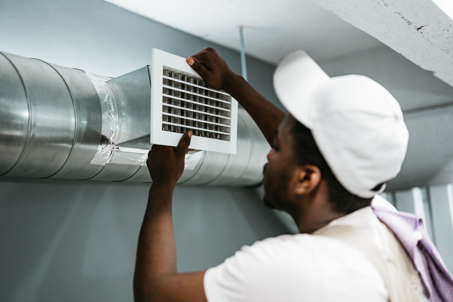 Person in white cap, replacing a vent cover on silver HVAC ductwork.