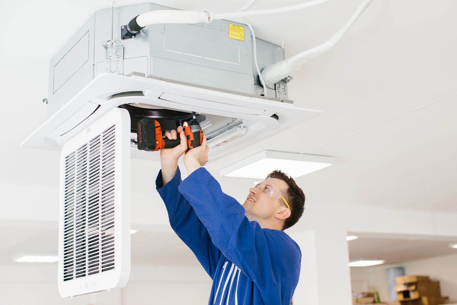 A person in blue overalls installs an air conditioning unit on a white ceiling, using a drill.