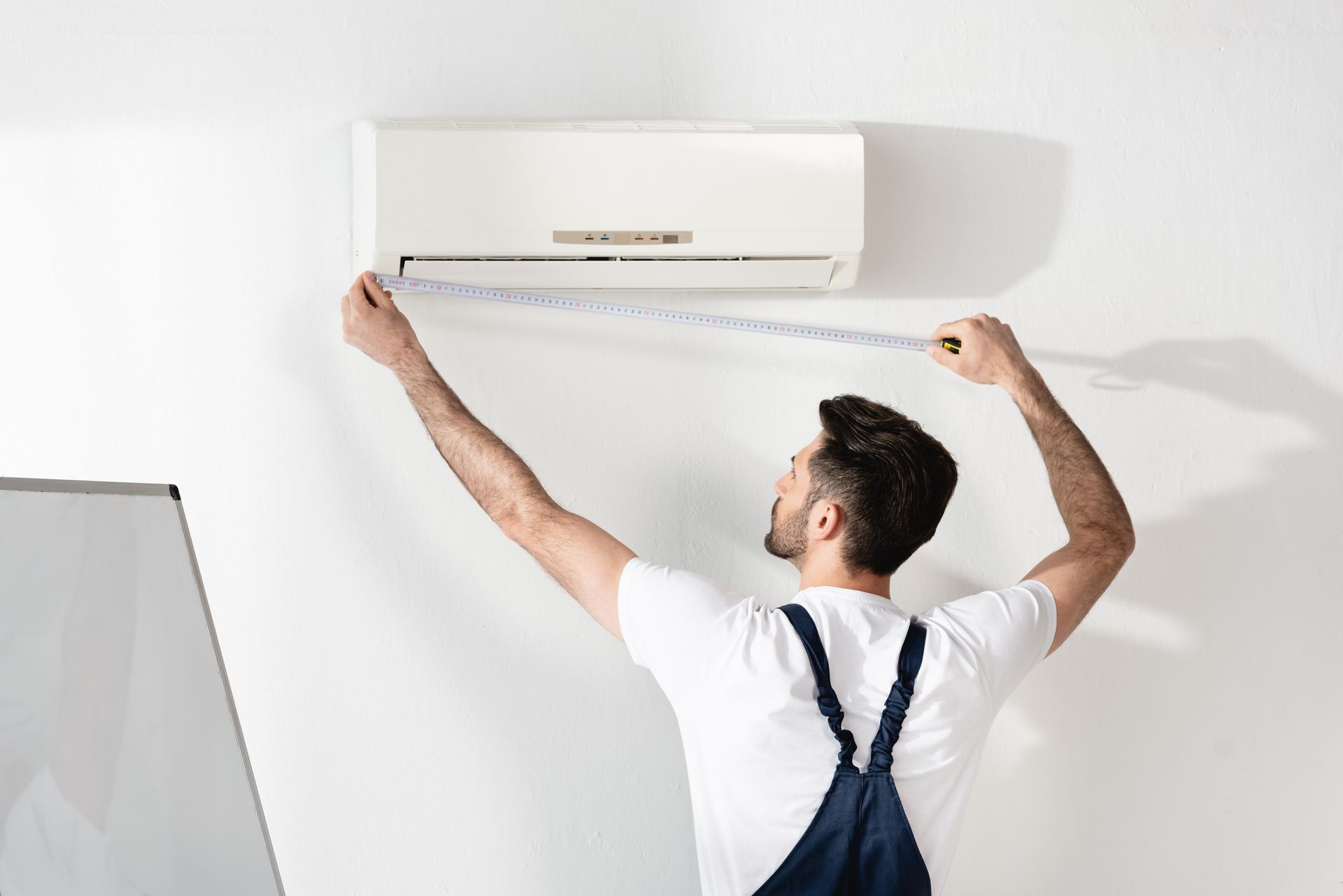 Man measures an air conditioner mounted on a white wall with a measuring tape.
