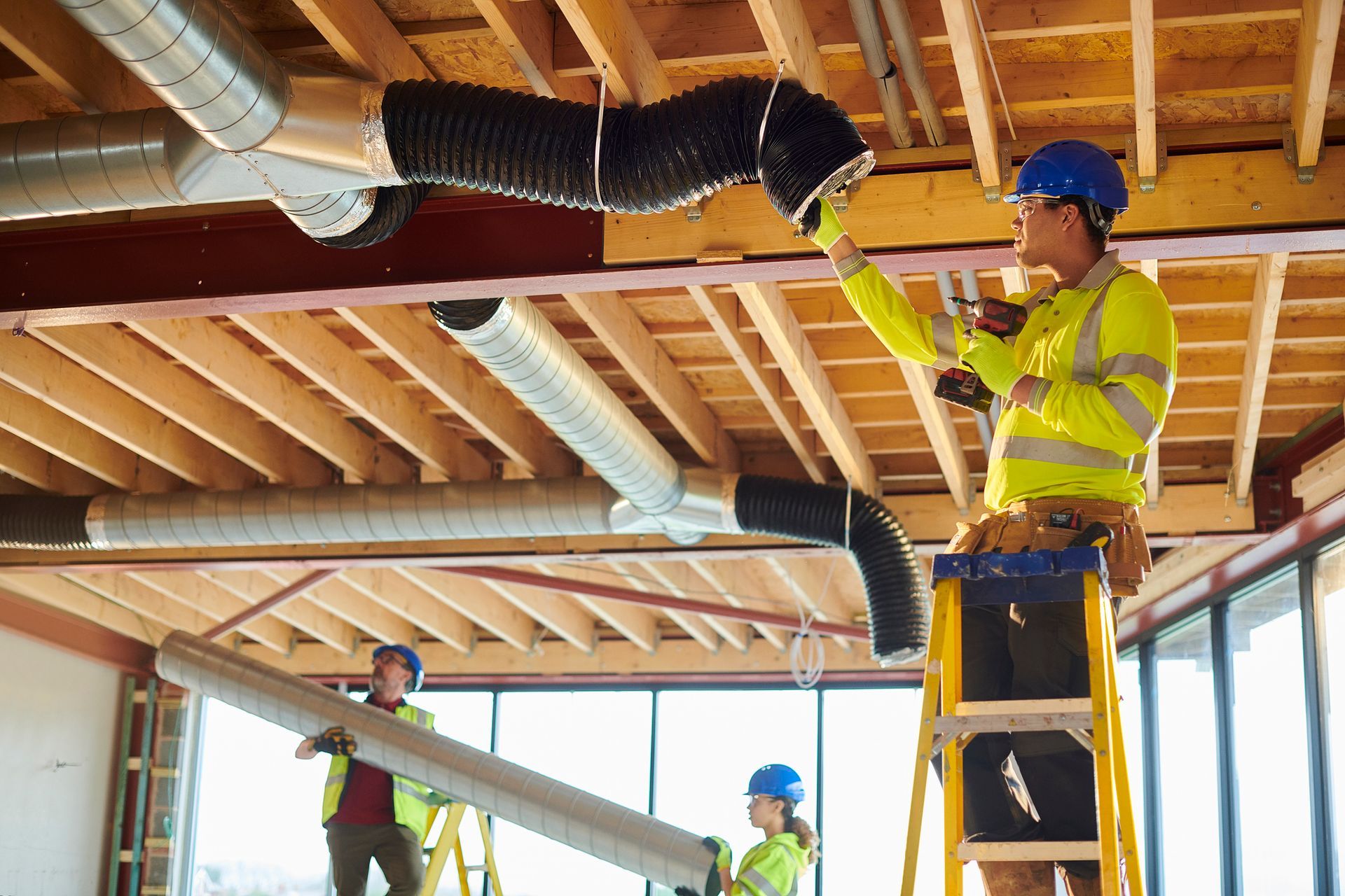 Construction workers installing HVAC ductwork in a building. They wear hard hats and safety vests.