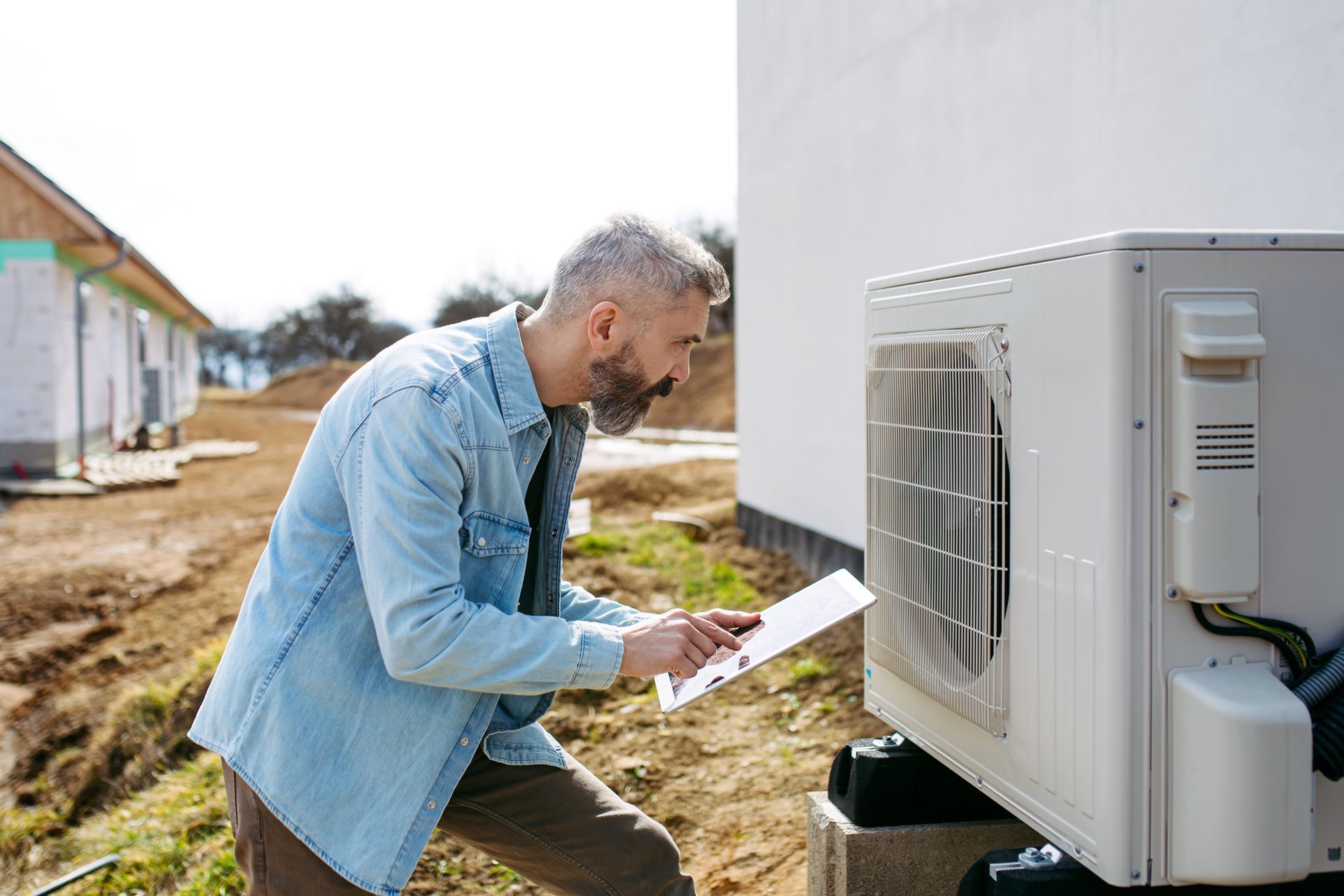 Man in denim shirt inspects outdoor AC unit, holding a clipboard, in a sunny yard.