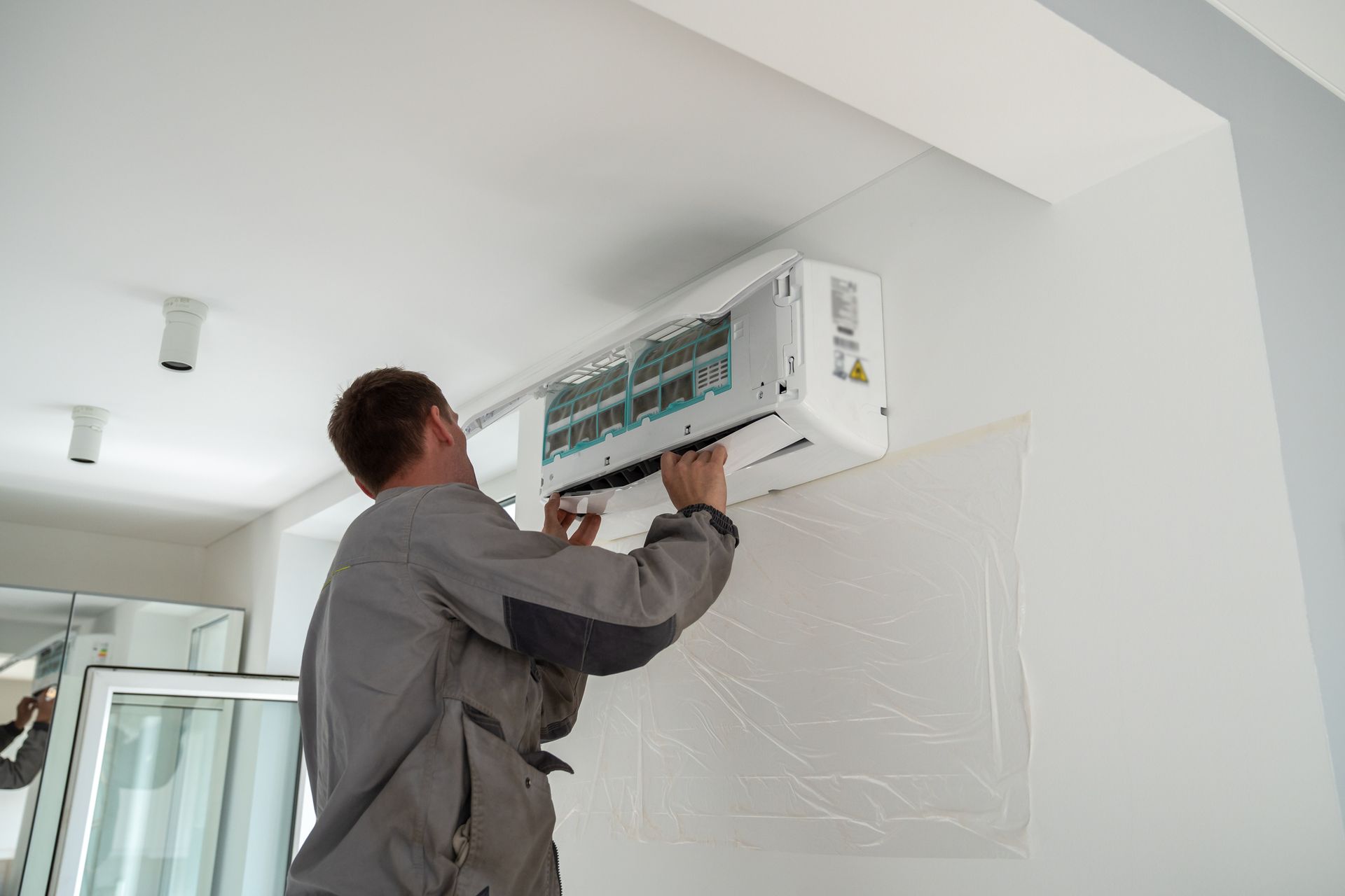 Man installing a wall-mounted air conditioning unit indoors, wearing a gray jumpsuit.