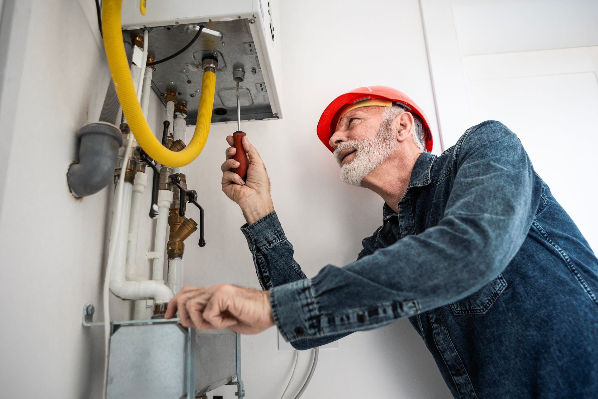 Handyman in a hard hat repairs a wall-mounted appliance with a screwdriver.