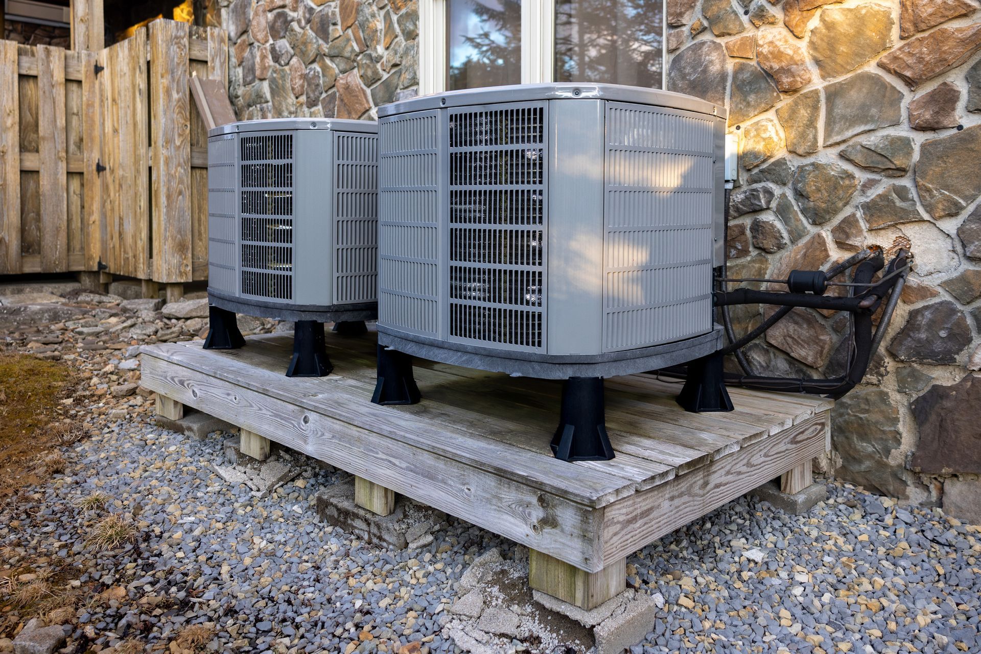 Two air conditioning units on a wooden platform against a stone wall.