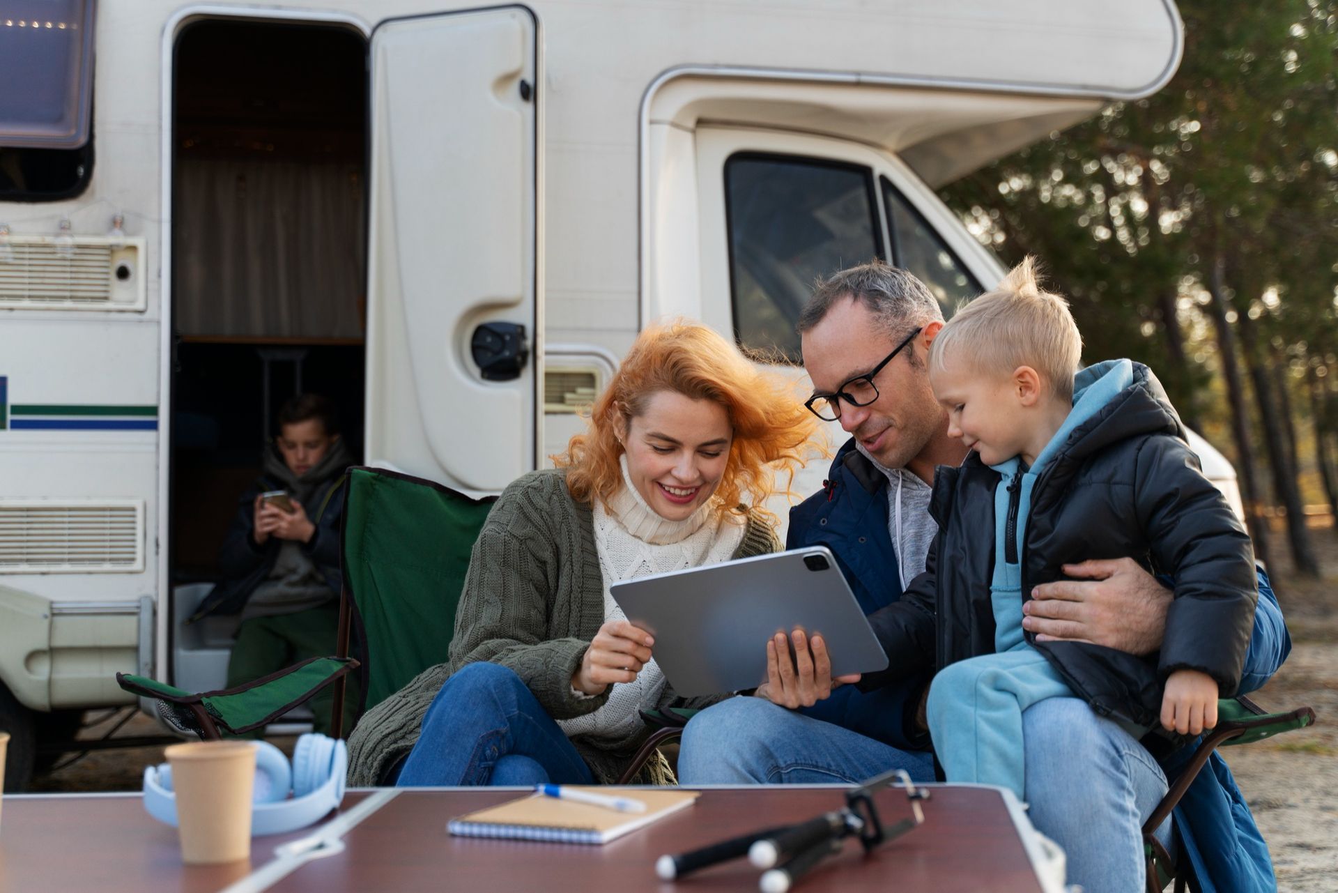 Friends outdoors by a camper, sharing a tablet at a table, with coffee cups and a relaxed campsite setting