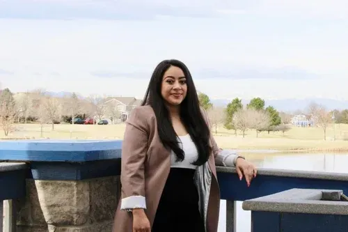Woman standing on a balcony with a blue railing, smiling in a beige coat outdoors.