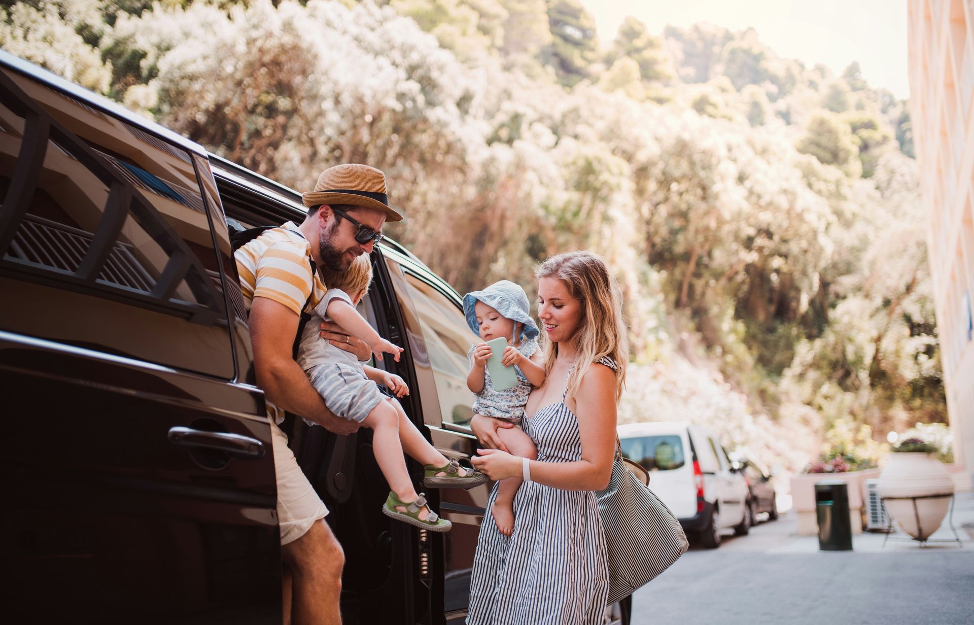 Family unloading children from a van on a sunny roadside, with bags and parked cars nearby.