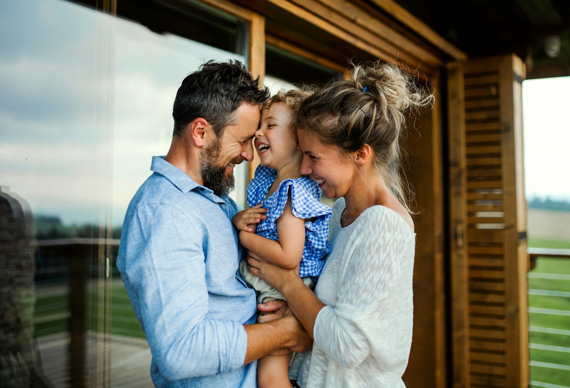 Family standing on a porch, smiling and hugging near a wooden house.