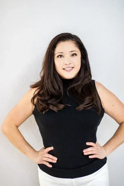 Woman with long dark hair wearing a black sleeveless top, standing with hands on hips against a light background.