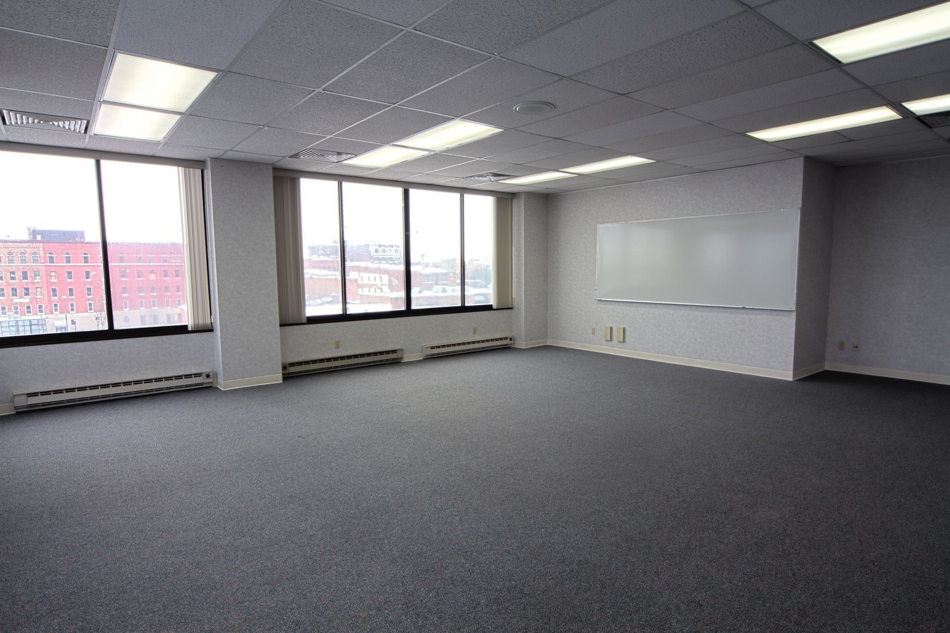 Empty office space with large windows and gray carpet. Whiteboard on wall.