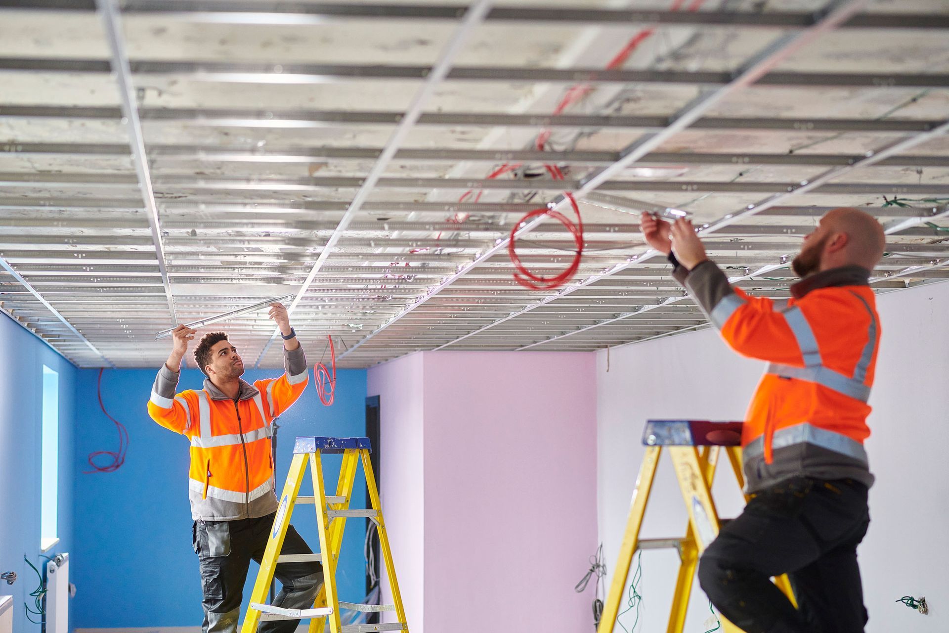 Two workers in orange vests install wires in a ceiling, using ladders, in a room with blue and pink walls.