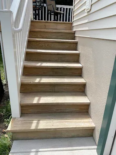 A set of wooden outdoor stairs with a white railing leading up to a deck beside a light-colored exterior wall.