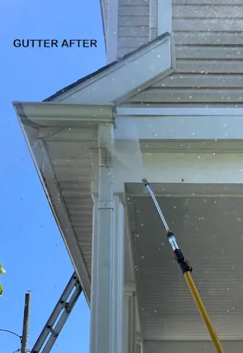 A yellow extension wand sprays water to clean a white house gutter under a clear blue sky.