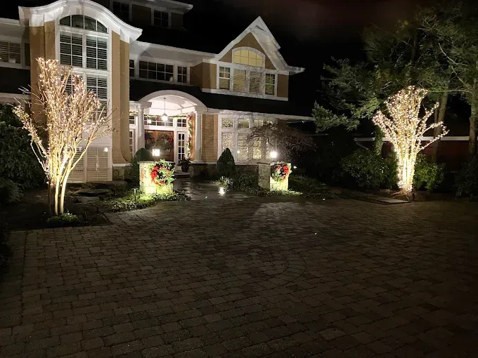 A large house at night with illuminated, light-wrapped trees and holiday wreaths decorating the stone driveway entrance.