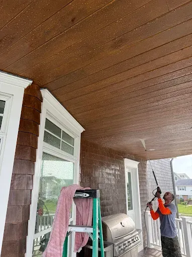 A worker uses a pressure washer to clean the wooden ceiling and siding of a house porch with a ladder and grill nearby.