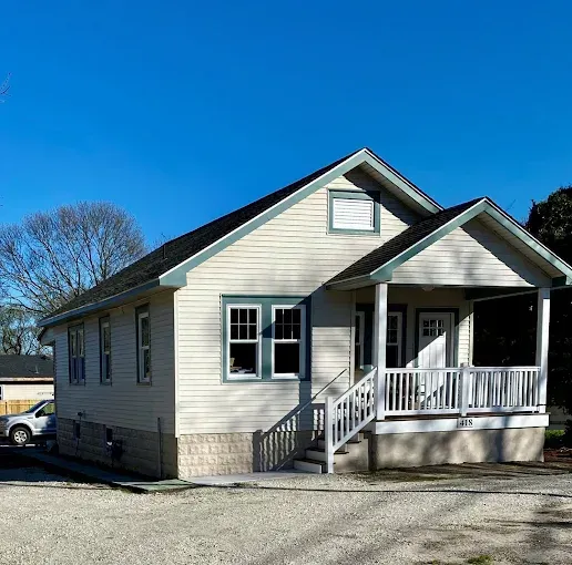 A small, one-story house with light beige siding, green trim, and a front porch, set under a clear blue sky.