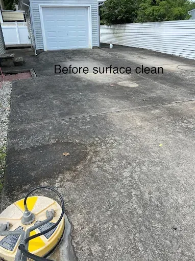 A yellow surface cleaner sits on a dark, stained concrete driveway in front of a garage. Text reads 