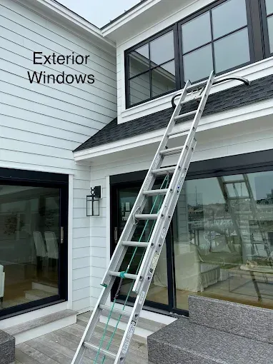 An aluminum extension ladder leans against the roofline of a white house, positioned to reach the upper exterior windows.