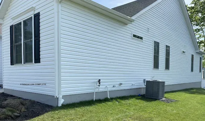 A side view of a white vinyl-sided house with black window shutters, a gray foundation, and an outdoor HVAC unit on grass.