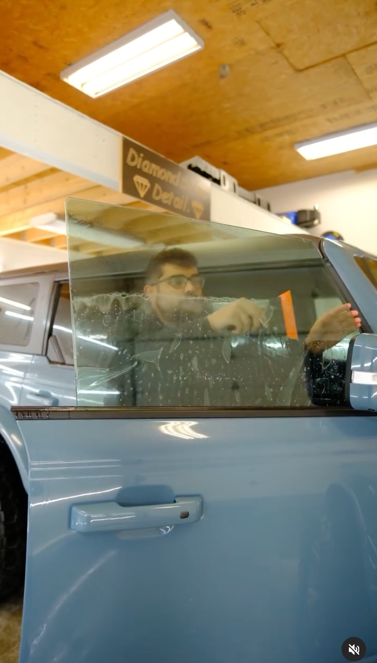 A person applying tint to a blue car window inside a shop. They use a tool to smooth the film.