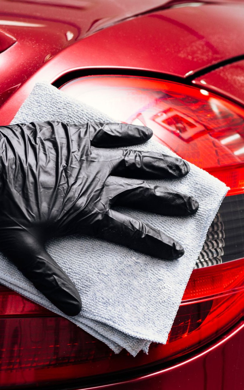 Black-gloved hand cleaning a red car's tail light with a microfiber cloth.