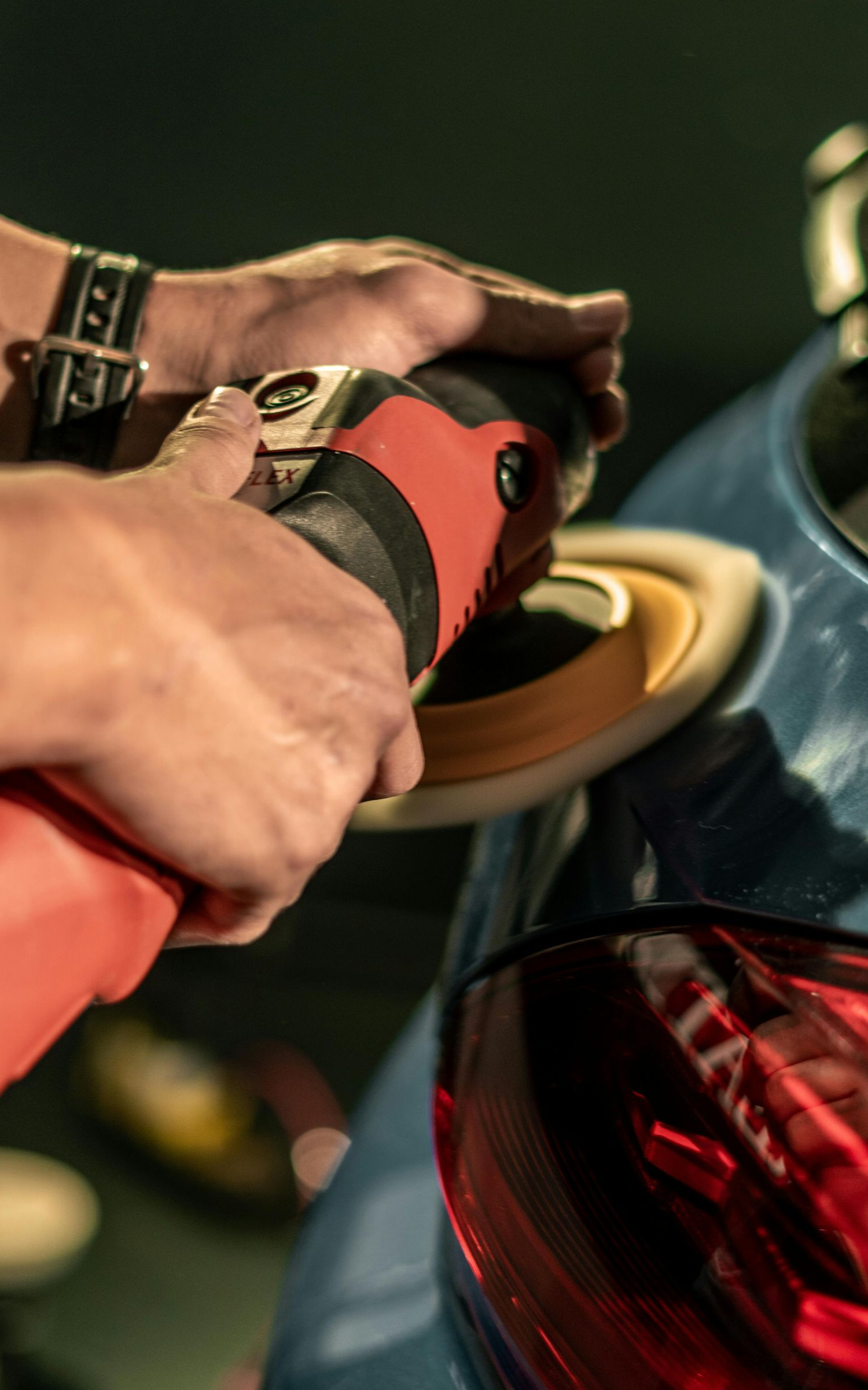 A person using a red and black power polisher on a blue car's rear panel.