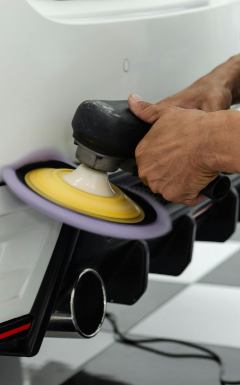 Person polishing a white car bumper with a power buffer, revealing the exhaust.
