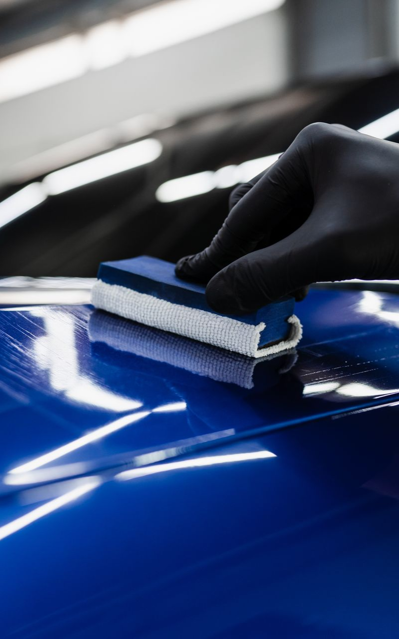 Gloved hand applying ceramic coating to a shiny blue car's surface with a blue applicator pad.