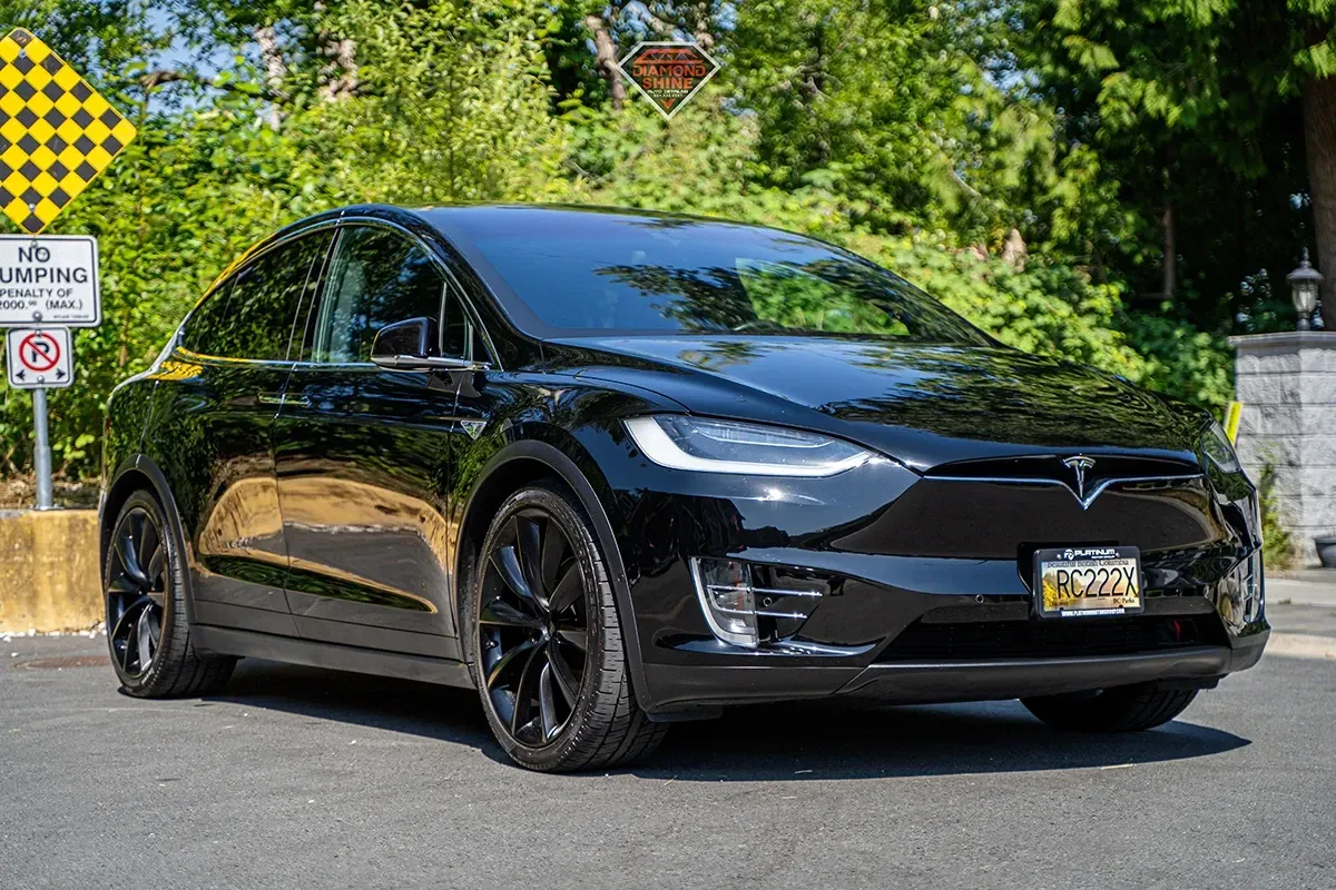 Black Tesla Model X parked on asphalt, with green foliage in background.