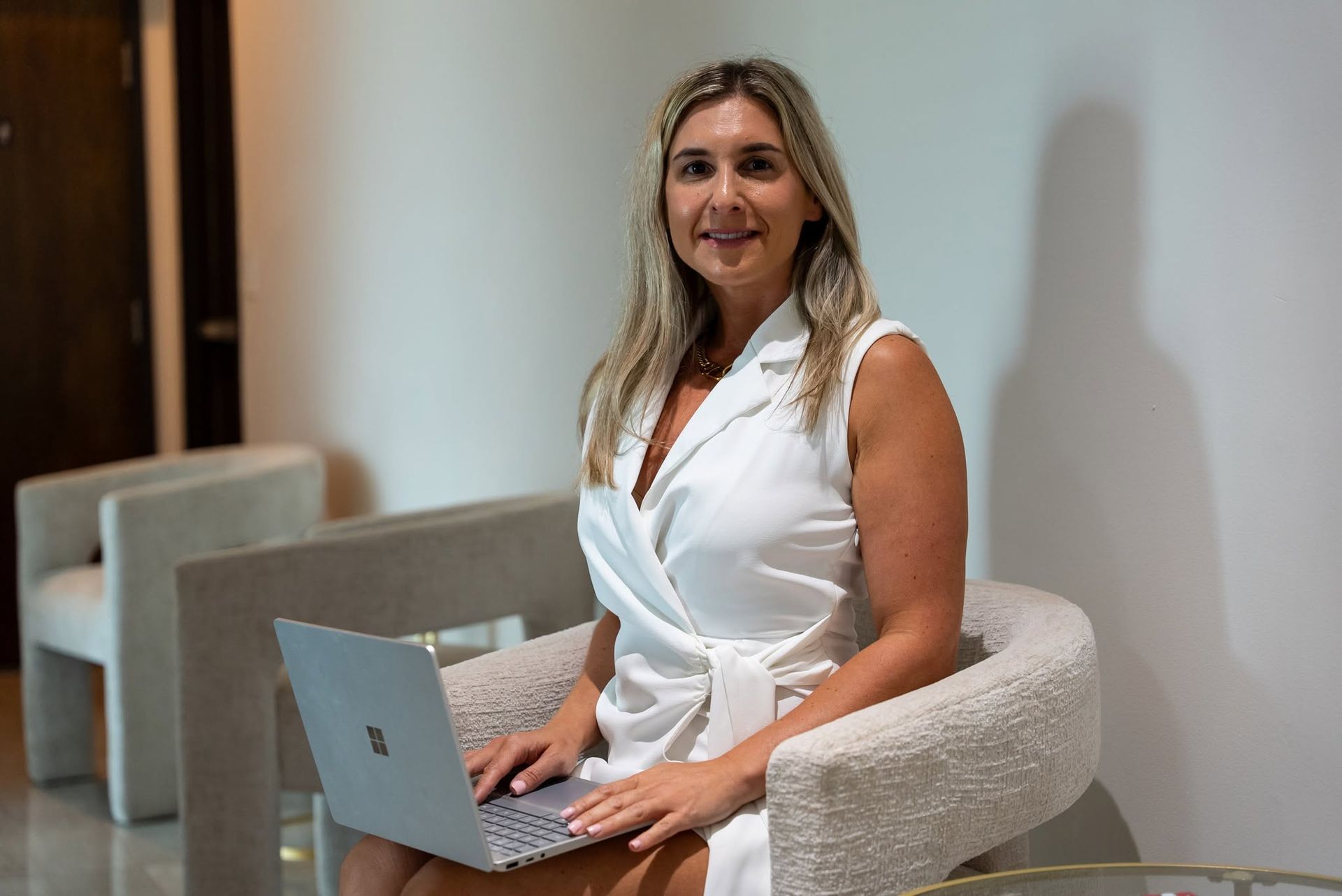 Woman sitting with a laptop in a bright modern lounge, smiling at the camera.
