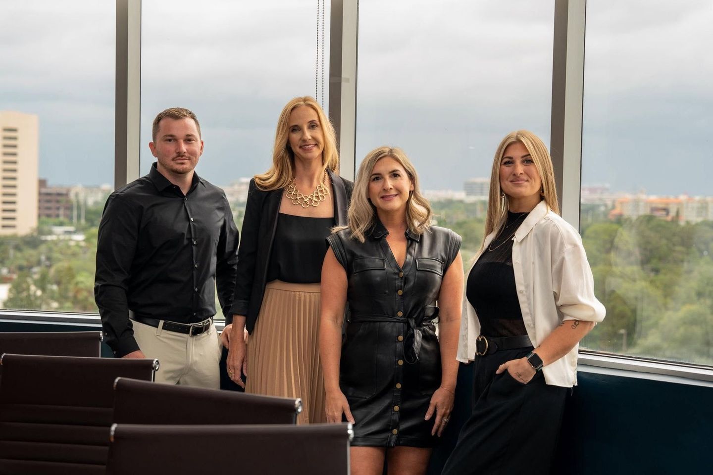 Four coworkers standing in an office conference room with large windows, dressed in business attire.