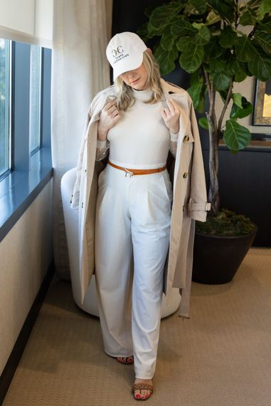 Woman in a beige coat and white cap standing indoors by a window and plant, wearing white pants and heels