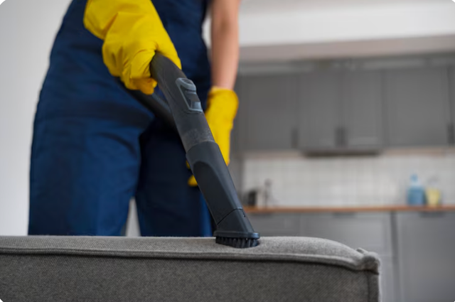 Person in yellow gloves vacuuming a gray couch in a kitchen.
