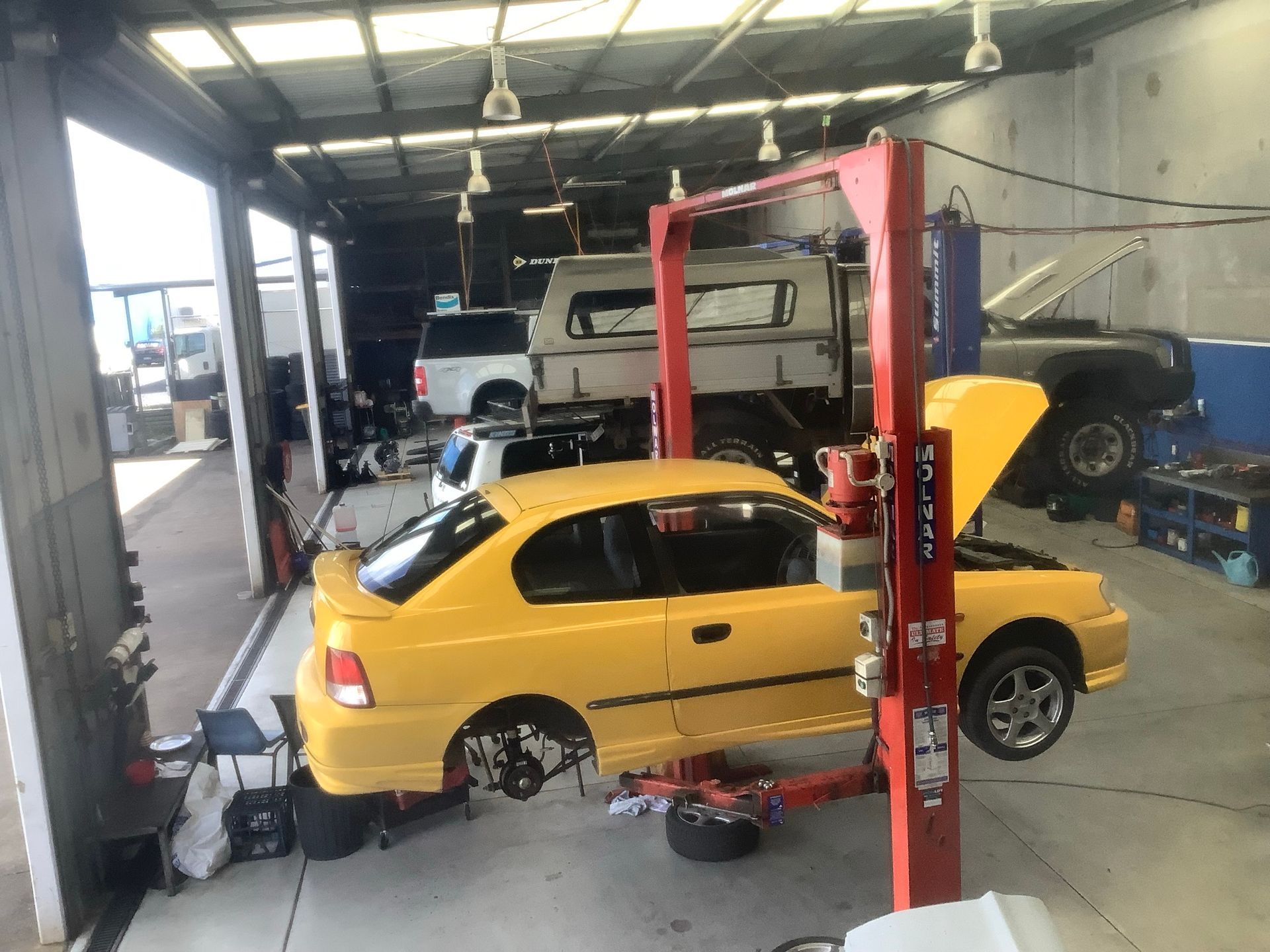 Yellow car on a lift in a mechanic's shop with hood open, other vehicles in the background — Pialba Brake Clutch & Steering Centre in Pialba, QLD