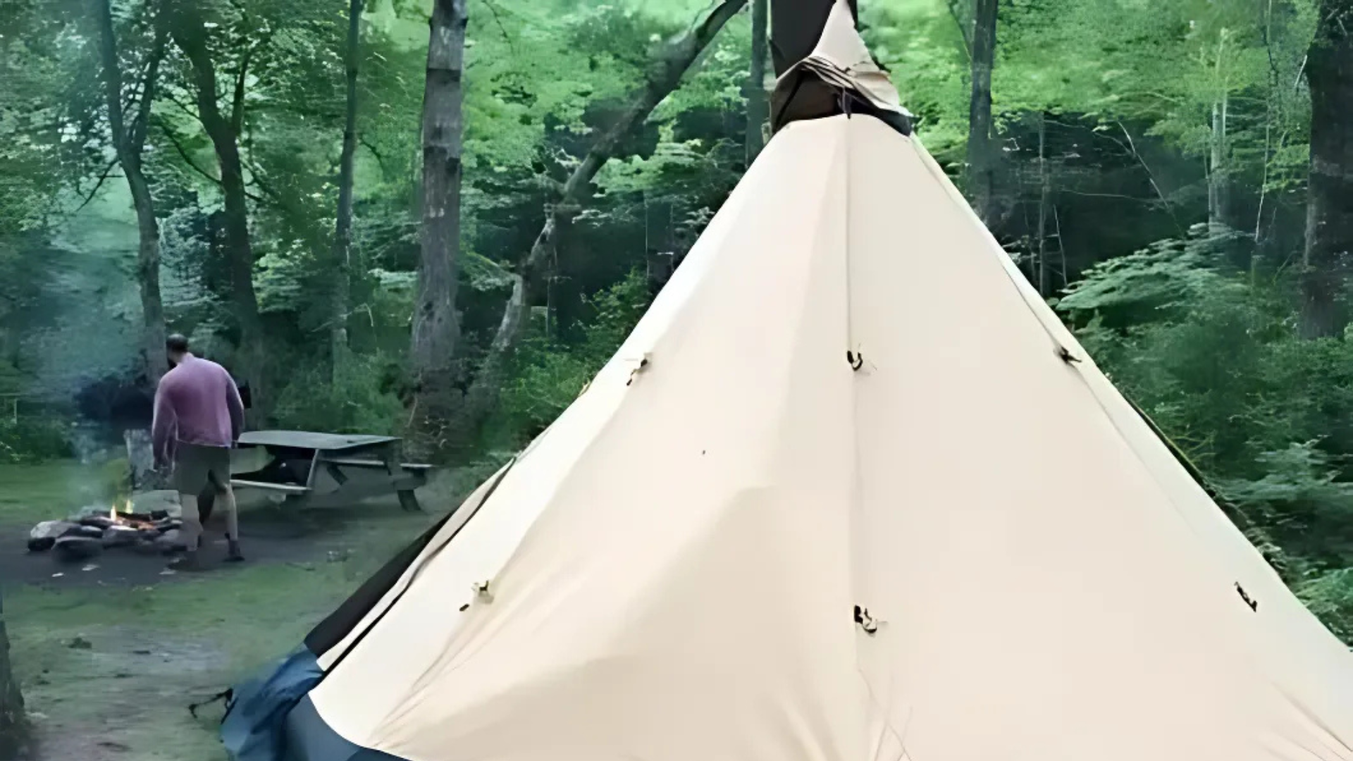 A man is standing in front of a teepee in the woods.