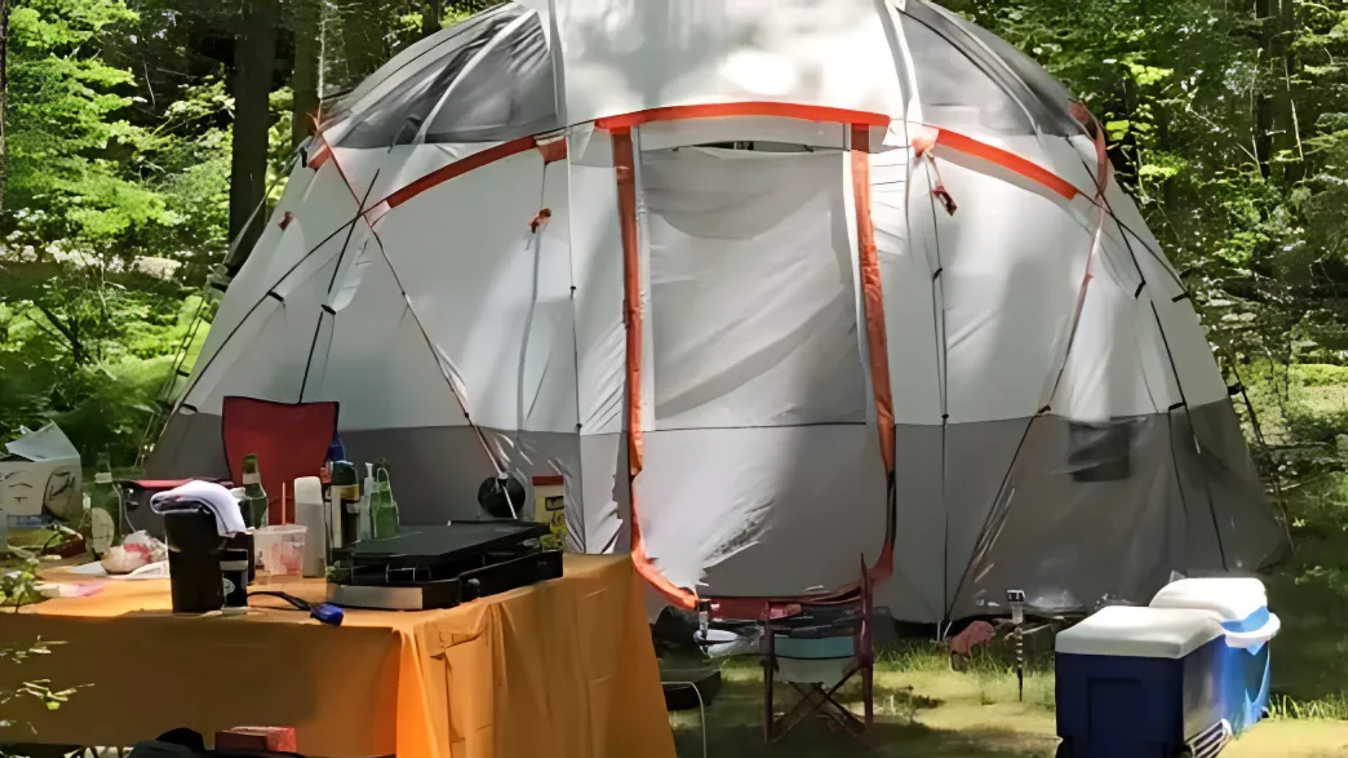 A large tent is sitting in the middle of a forest next to a table and chairs.