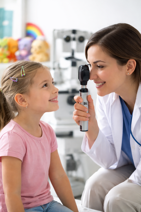 An eye doctor holds an ophthalmoscope to examine a child’s eye in an exam room.