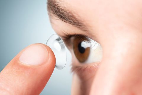 A close-up view of a person’s finger holding a clear contact lens in front of an open brown eye.