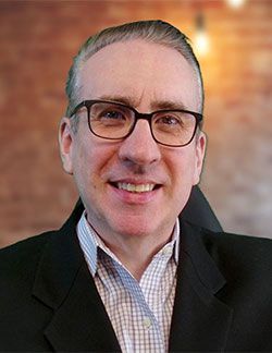 A man wearing glasses and a suit is smiling for the camera.