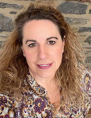 A woman with long curly hair is smiling for the camera in front of a stone wall.