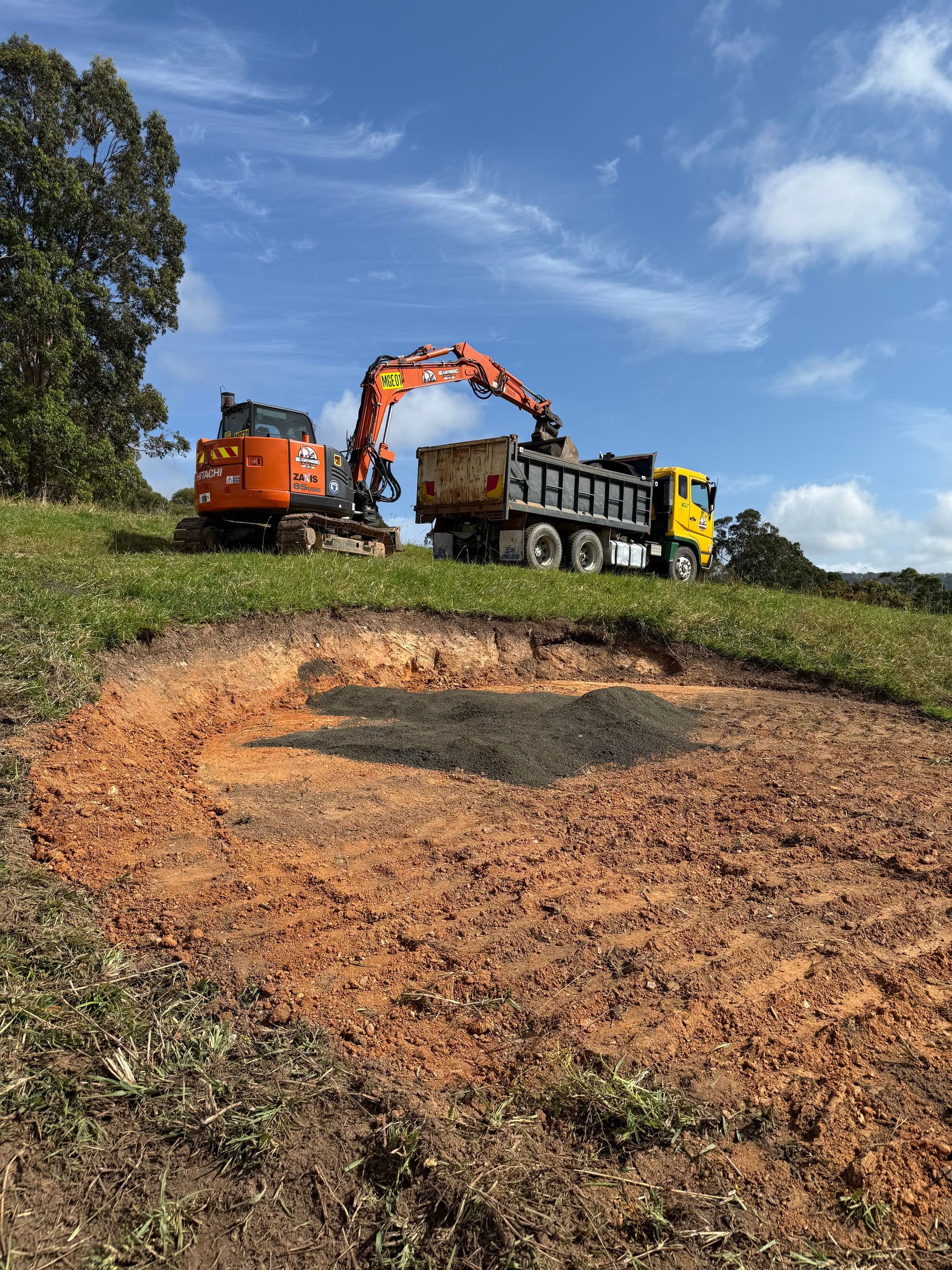 Excavator loading dirt into a dump truck on a grassy hill; blue sky background.