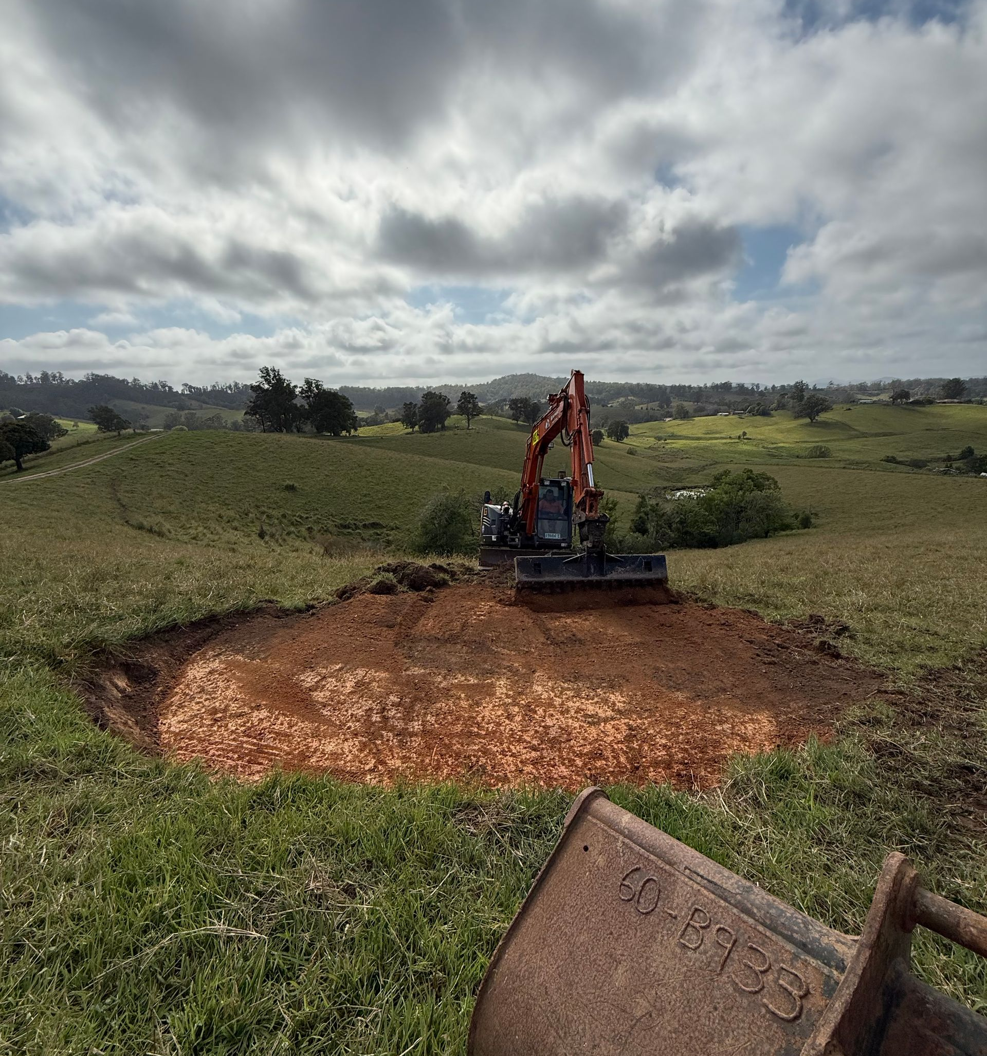 Excavator digging in a grassy field under a cloudy sky. A bucket is in the foreground.