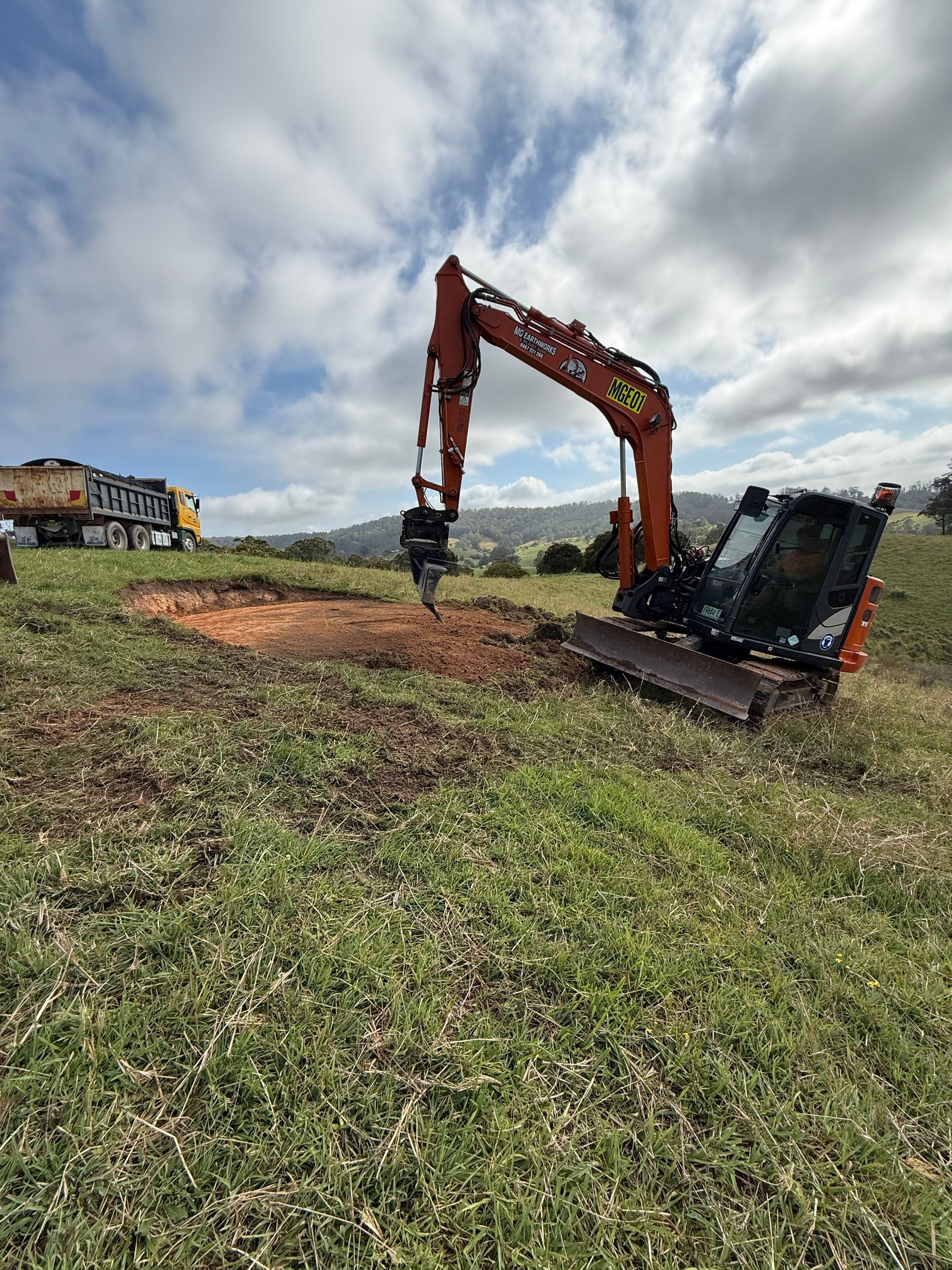 Orange excavator digging into a grassy field on a sunny day. A dump truck sits in the background.