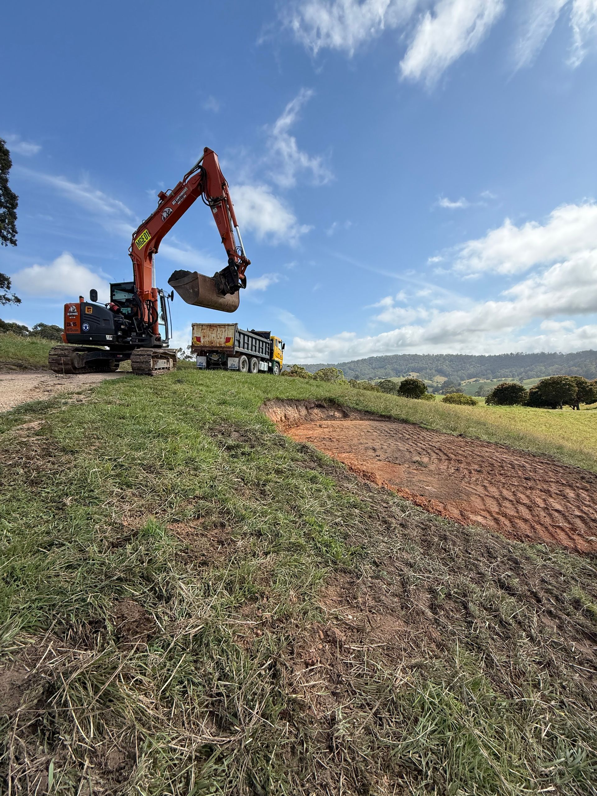 An excavator loading dirt into a dump truck on a grassy hillside under a blue sky.