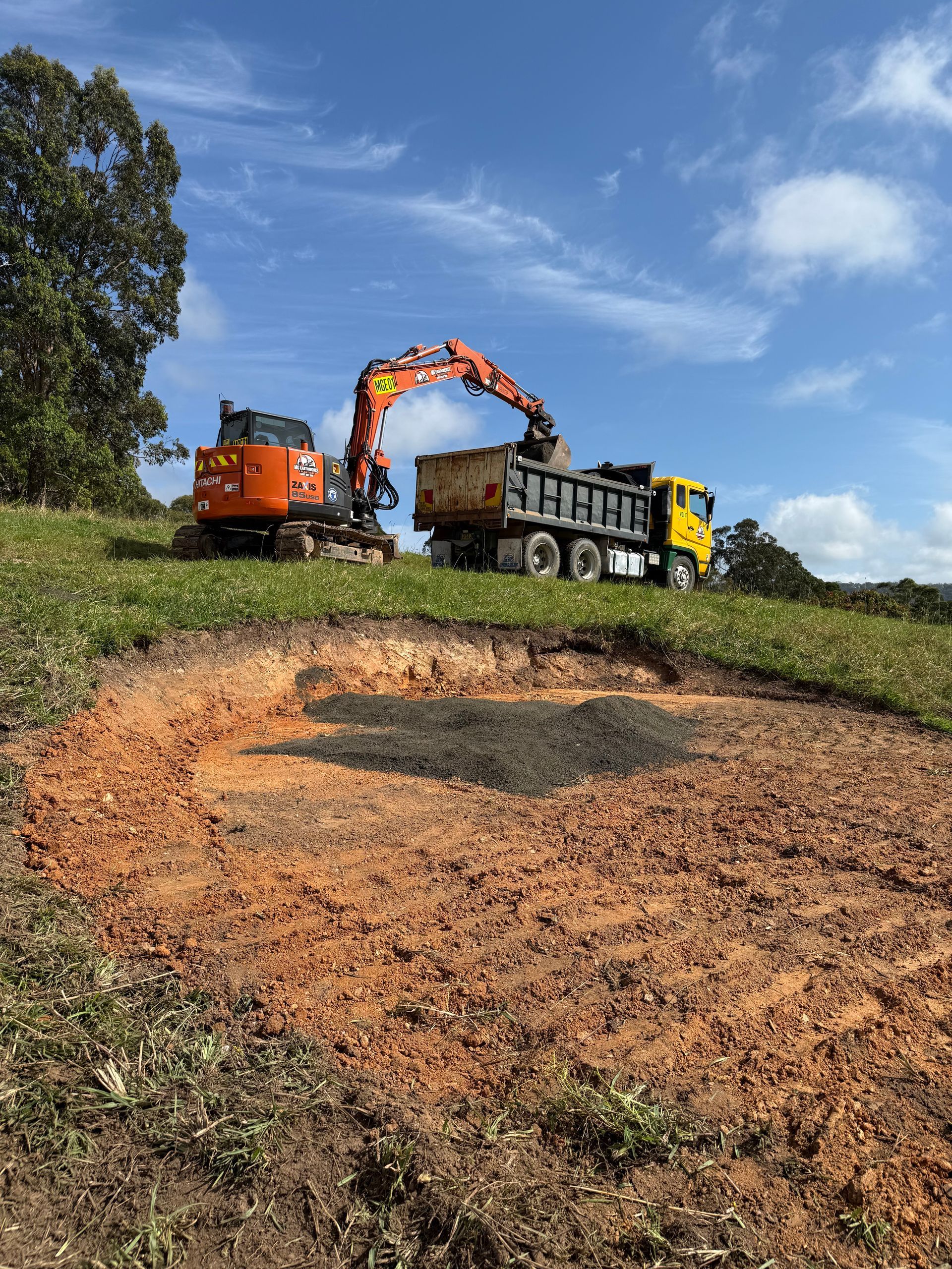 Orange excavator loading dirt into a yellow dump truck on a grassy hill; blue sky.
